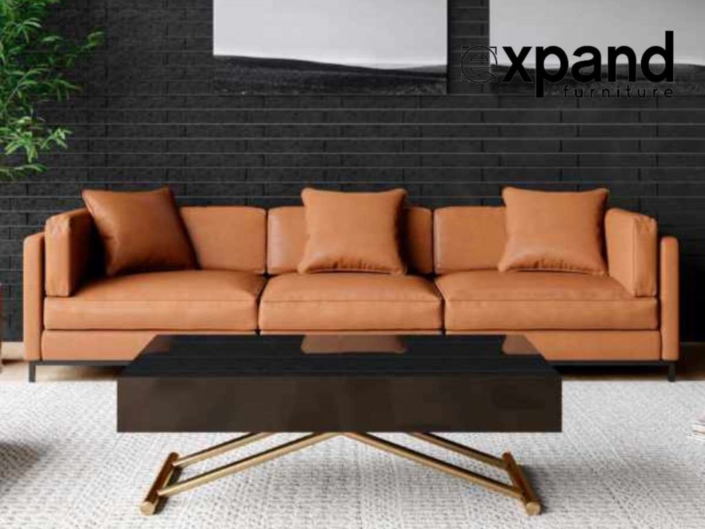 Modern living room with a tan leather sofa and a glass box coffee table on a patterned rug against a black brick wall