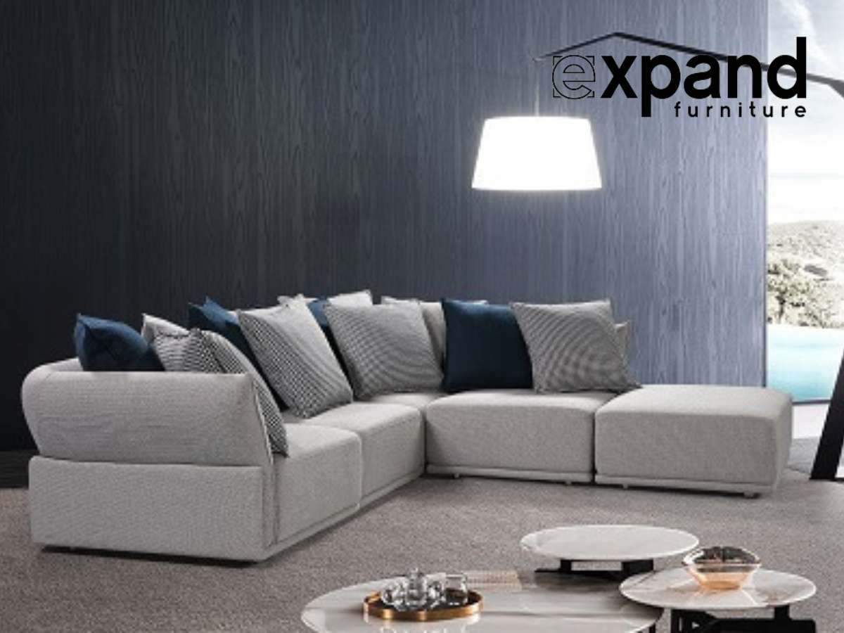 A contemporary living room featuring a modular sectional sofa with variously sized cushions in shades of gray and blue, set against a dark textured wall with a large pendant lamp overhead and a round multi-tiered coffee table in the foreground.